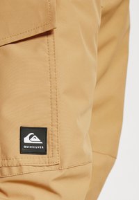 Pantalon couleur kaki avec un écusson rectangulaire noir du logo Quiksilver près d'un rabat de poche latérale cousu.