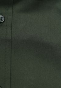 Donkergroene stof met een gladde textuur, voorzien van een knoopdetail en contrasterende stiksels langs de naad.