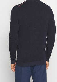 Tummansininen pitkähihaisten teksturoidun villapaidan, jossa on ribbitut hihat ja helma, ja olkapäällä kapea punainen korostus.