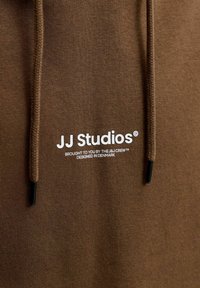 Tissu de sweat à capuche marron avec logo texte blanc "JJ Studios" et deux cordons marron avec embouts noirs visibles.