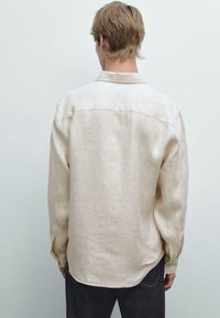 Beige linen shirt with a relaxed fit, long sleeves, and a classic collar. Smooth texture, curved hem, and buttoned cuffs visible from the back.