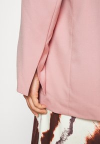 Veste rose clair cintrée en tissu lisse, avec des fentes latérales et des manches ajustées. Main posée sur une jupe à motifs.