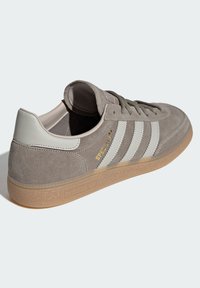 Γκρι σουέντ αθλητικό παπούτσι με λευκές δερμάτινες λεπτομέρειες και τρεις ρίγες. Σόλα από λαστιχένιο υλικό καραμέλας. Διακοσμημένο με χρυσό σήμα στην πλευρά και υφασμάτινη υφή.