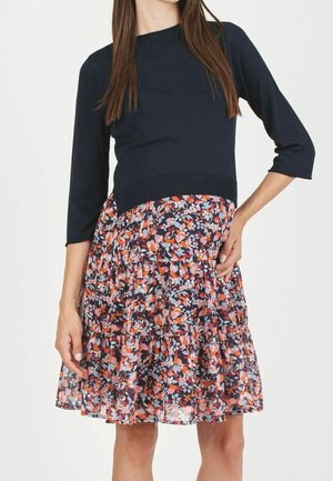 Femme portant un haut bleu marine à manches 3/4 associé à une jupe fleurie longueur genou aux tons orange, rose et bleu clair.