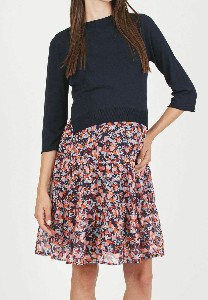Femme portant un haut bleu marine à manches 3/4 associé à une jupe fleurie longueur genou aux tons orange, rose et bleu clair.