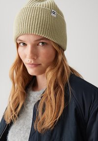 Light green ribbed knit beanie with a small logo tag. Model has wavy hair and is wearing a gray sweater under a dark jacket.