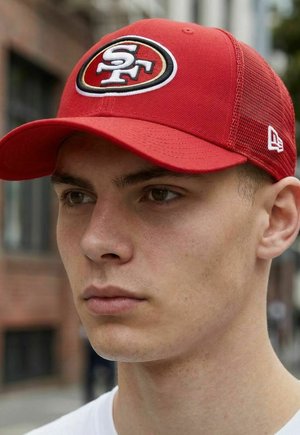 Young man wearing a red mesh baseball cap with San Francisco 49ers logo, standing outdoors in an urban setting.