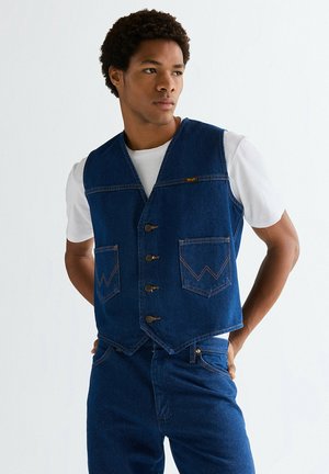 Mand iført en hvid t-shirt og blå denimvest og jeans, stående med hænderne bag ryggen, kiggende til siden.