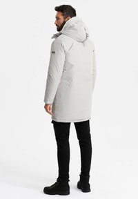 Ljusgrå vadderad parkas med huva, med en dragkedja fram, justerbara dragsko och en slät textur. Kombinerad med svarta byxor och stövlar.
