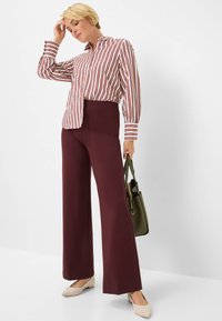 Blusa a righe rossa e bianca, abbinata a pantaloni larghi bordeaux. La modella tiene una borsa verde e indossa delle scarpe basse chiare.
