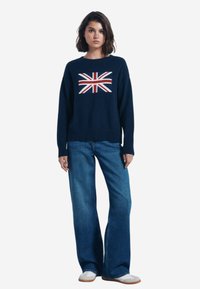 Maglione lavorato a maglia blu navy con design della Union Jack sulla parte anteriore, abbinato a jeans a gamba larga blu chiaro e scarpe beige slip-on.