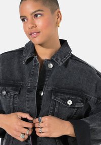 Schwarze Jeansjacke mit silberner Hardware, Brusttaschen und Kragen. Das Modell hält die Jacke und zeigt Ringe an beiden Händen.