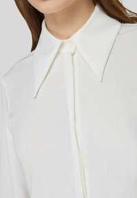 Blusa bianca con un ampio colletto a punta, chiusura a bottoni e una texture di tessuto liscia e leggermente lucida. Cuciture dettagliate visibili.