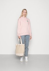 Felpa rosa con tasca frontale e ricamo floreale, abbinata a jeans strappati blu chiaro e una borsa a tracolla beige a righe. Sneakers bianche.