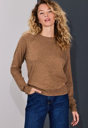Blondynka o kręconych włosach, ubrana w brązowy sweter w cętki i niebieskie dżinsy, pozuje z jedną ręką w kieszeni na dwutonowym szarym tle.