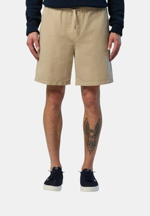Shorts in cotone beige con elastico in vita e cordoncino. Lunghezza corta, texture liscia, abbinati a sneakers scure e una gamba tatuata.