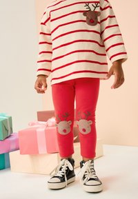 Leggings rosse con applicazione di renna sulle ginocchia, abbinate a una maglia a maniche lunghe color crema con strisce rosse e una tasca sul petto con renna. Scarpe da ginnastica nere.