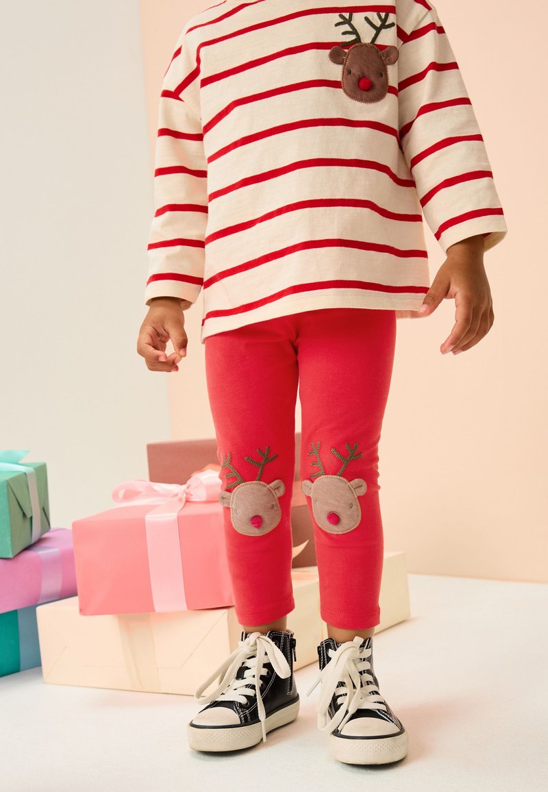 Leggings rouges avec appliqué de renne sur les genoux, associés à un t-shirt à manches longues crème avec des rayures rouges et une poche poitrine en forme de renne. Baskets noires.
