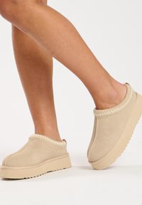 Beige slip-on shoes with a suede finish, textured collar, and a thick, ridged sole. Subtle stitching detail around the upper.