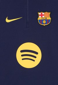 Polo-shirt bleu marine en matière synthétique, arborant un logo Nike doré, un écusson multicolore du FC Barcelone et un logo Spotify jaune.