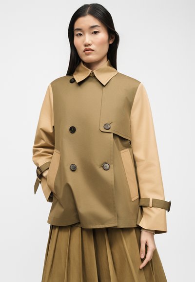 Γυναίκα που φοράει δίχρωμο καφέ καμπαρντίνα με λεπτομέρειες κουμπιών και φούστα με πιέτες, στέκεται μπροστά σε απλό φωτεινό φόντο.