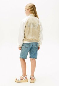 Niño con cabello rubio que lleva una chaqueta beige y blanca, pantalones cortos de mezclilla y sandalias blancas, de pie de espaldas sobre un fondo blanco.