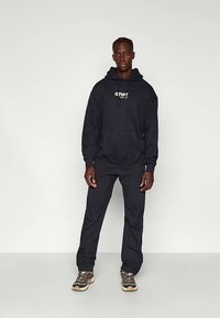 Νέος άνδρας στέλνει πρόσωπο φορώντας μαύρη κουκούλα με λευκό λογότυπο, μαύρο παντελόνι και μπεζ αθλητικά παπούτσια μπροστά σε απλό ανοιχτό φόντο.