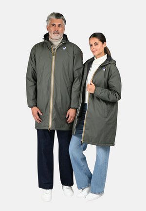 Vestes longueur genou de couleur vert olive avec des fermetures éclair jaunes contrastantes. Les deux ont des capuches et des poches latérales, portées sur des pulls à col roulé clairs et des jeans.