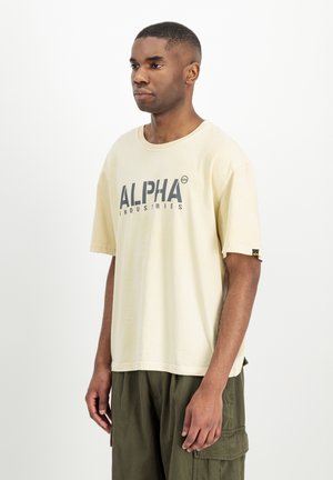 Čovjek stoji, nosi bež Alpha Industries majicu i maslinasto zelene cargo hlače na jednostavnoj bijeloj pozadini.