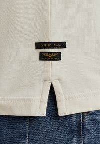 Cremefarvet bomulds T-shirt med split nederst i siden. Har to sorte mærker med hvid og gul tekst, der beskriver typenummer og mærke.