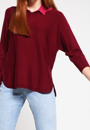 Femme portant un pull ample rouge foncé par-dessus une chemise à col rouge vif avec un jean bleu clair, debout devant un fond blanc uni.