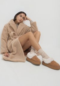 Frau, die auf dem Boden sitzt, trägt einen beigen Teddy-Mantel, passende Socken und braune Hausschuhe mit Lammfellfutter auf einer Plattform, mit der Hand im Nacken.