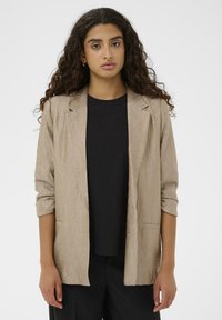 Blazer beige texturé avec une coupe décontractée, fermeture à un bouton, et manches retroussées, porté sur un t-shirt noir.