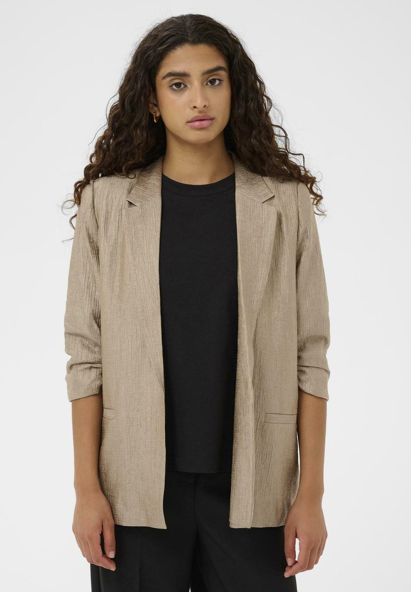 Blazer beige texturé avec une coupe décontractée, fermeture à un bouton, et manches retroussées, porté sur un t-shirt noir.