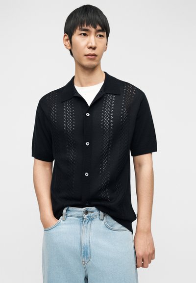 Νεαρός άνδρας που φοράει μαύρο πουκάμισο με κοντά μανίκια και κάθετες υφασμάτινες λεπτομέρειες πάνω από ένα λευκό εσώρουχο και ανοιχτόχρωμα τζιν παντελόνια.