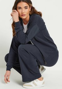 Marineblauwe ritsjack en bijpassende sweatpants van zacht materiaal. Model hurkt met een hand op haar knie, met witte sneakers aan.