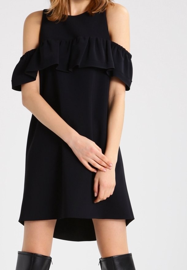 Woman wearing a black cold-shoulder dress with ruffle detail and black knee-high boots, standing against a plain light background.