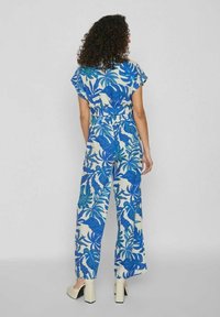 Květinový overal má modro-zelené listové vzory na krémovém pozadí. Má krátké rukávy, stahovaný pas a široké nohavice.