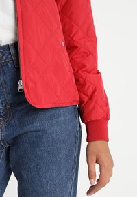 Veste matelassée rouge avec fermeture éclair et boutons-pression portée sur un jean bleu, montrant le bras gauche et la main contre un fond blanc uni.