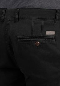 Pantalons noirs avec une texture lisse, dotés d'une poche arrière fermée par un bouton marron et d'une étiquette de marque. Design simple et ajusté.