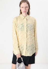 Camicia di un delicato giallo chiaro con una trama sfrangiata, chiusura frontale con bottoni e colletto a punta, abbinata a una gonna nera e a una borsa argentata.