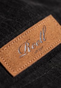 Tela negra con acabado texturizado presenta un parche rectangular de cuero marrón, grabado con la palabra "Reell" en letras plateadas.