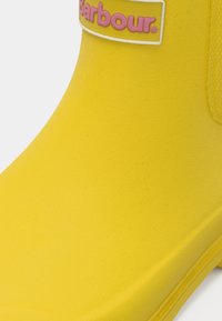 Botte en caoutchouc jaune avec une surface texturée et un patch logo rose. Design bas, bout arrondi et bords lisses.