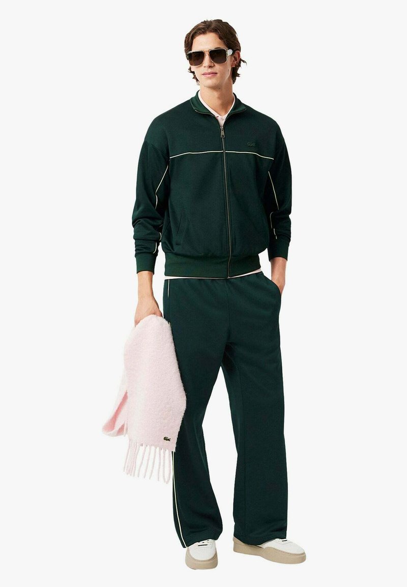 Tuta verde scuro con dettagli bianchi, giacca con cerniera, pantaloni a vestibilità morbida e sciarpa rosa chiaro. Include sneakers bianche e occhiali da sole.