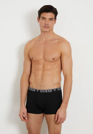 Boxeri negri până la talie cu un elastic alb cu logo-ul "GUESS". Realizați dintr-un material moale, cu o croială ajustată și o textură netedă. Modelul stă fără tricou.