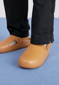 Brune gummiklipper med utskårne sider, teksturerte såler og justerbare stropper. Brukt med svarte bukser på en lyseblå overflate.