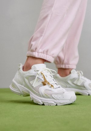 Białe siateczkowe i skórzane sneakersy ze złotym łańcuszkiem, noszone z jasnoróżowymi spodenkami dresowymi na zielonej podłodze.