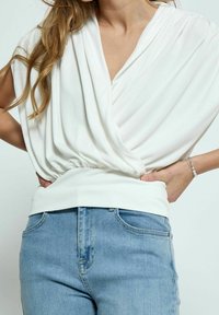 Blusa blanca drapeada con un escote en V profundo, cintura fruncida y textura suave, combinada con vaqueros de mezclilla azul claro con bolsillos clásicos.