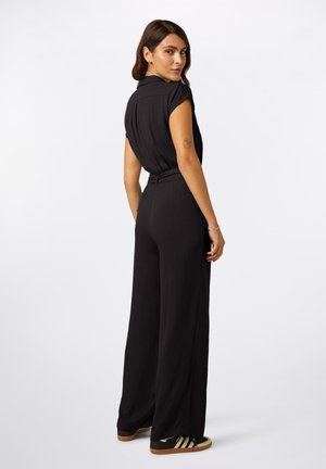 Vrouw met bruin haar, gekleed in een zwarte mouwloze blouse en zwarte broek met wijd uitlopende pijpen, kijkt over haar schouder terug, staand tegen een witte achtergrond.