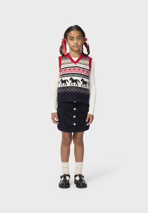 Ragazza giovane con capelli intrecciati e nastri rossi che indossa un maglione senza maniche con cavalli e cuori, camicia bianca a maniche lunghe, gonna blu navy e scarpe nere.